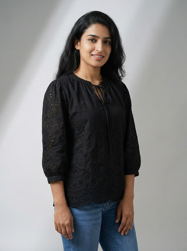 Woman wearing a black eyelet lace blouse with 3/4 sleeves and tie-neck, styled with blue jeans in a studio setting.